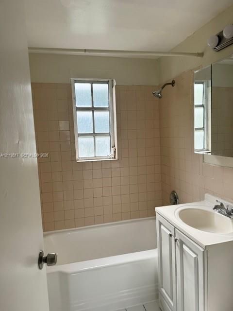 Bathroom featuring a white vanity cabinet with a drop-in sink, tiled walls, a standard bathtub, a multi-pane window, and a wall-mounted light fixture