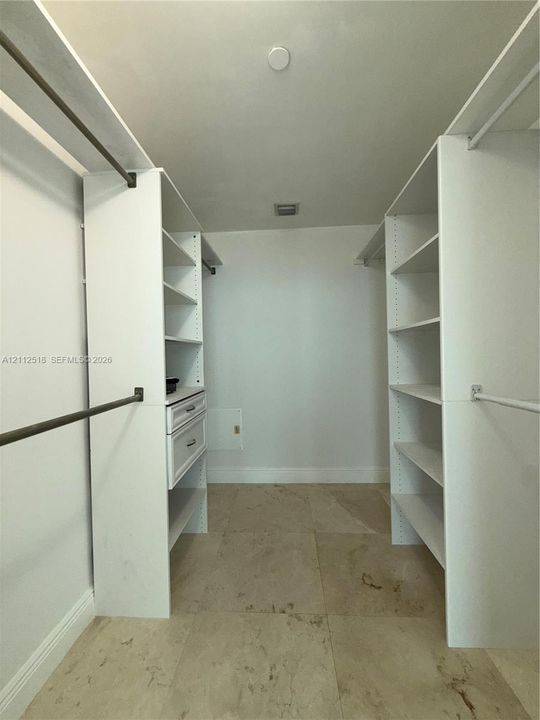 Walk-in closet featuring white built-in shelving, integrated dresser drawers, metal hanging rods, and stone-finish tile flooring
