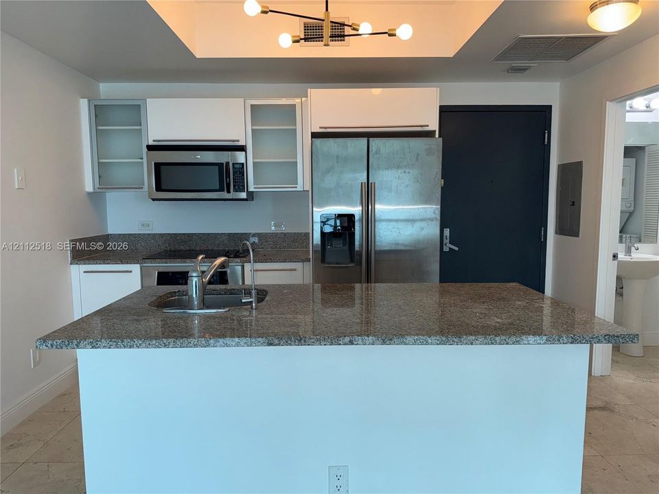 Kitchen featuring stone-surface countertops, stainless steel appliances, white cabinetry with glass-front uppers, a central island with an integrated sink, and modern pendant lighting