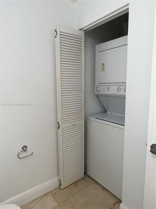 Laundry closet featuring a stackable washer and dryer unit, louvered bi-fold door, and light-toned tile flooring