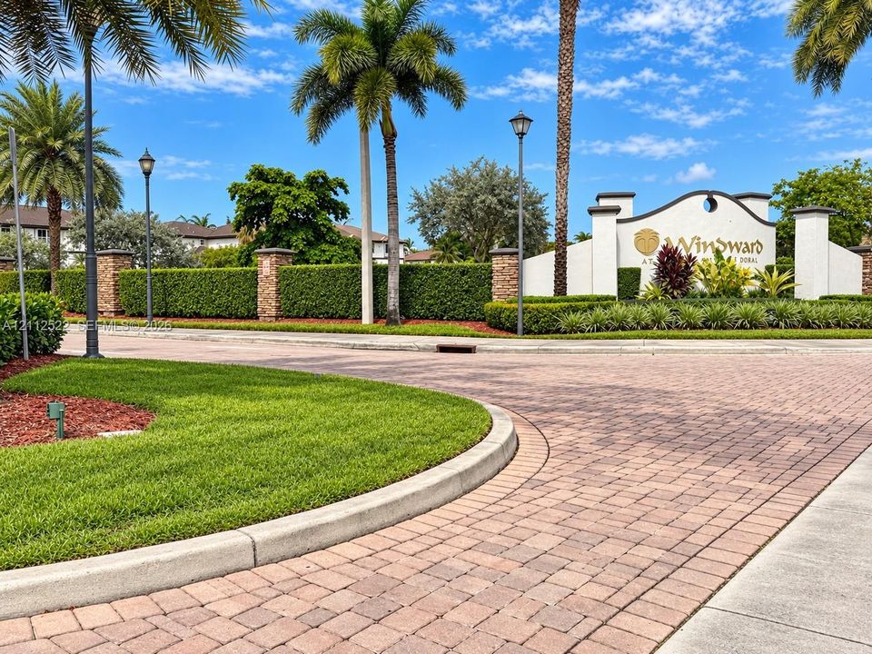 Gated community entrance featuring decorative brick paving, stone-accented masonry pillars, mature palm landscaping, and manicured hedges