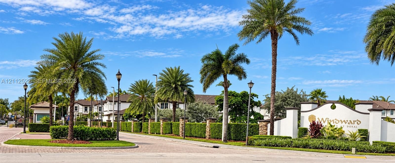 Gated entrance featuring masonry perimeter walls, manicured perimeter hedges, mature palm trees, and stone-paver driveway