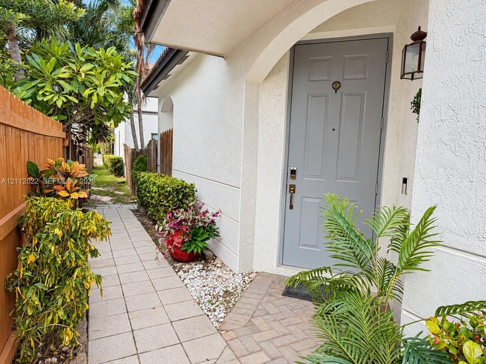 Entryway featuring a solid-panel gray front door, stucco exterior finish, arched architectural detail, decorative exterior wall-mounted lantern, and a herringbone-patterned paver landing