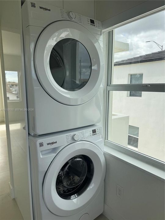 Stacked Whirlpool washer and dryer unit featuring digital controls and front-loading glass doors, positioned near a large window with white-frame casing