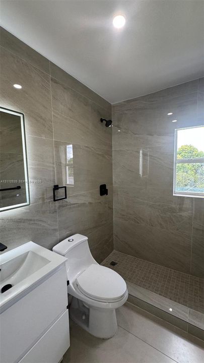 Large-format stone-style wall tiling, walk-in shower with mosaic tile flooring, contemporary white vanity, backlit mirror, and matte black hardware