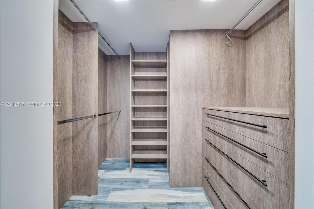 Walk-in closet featuring custom wood-finish cabinetry, floor-to-ceiling shelving, built-in dresser drawers, matte black hardware, and wood-finish flooring