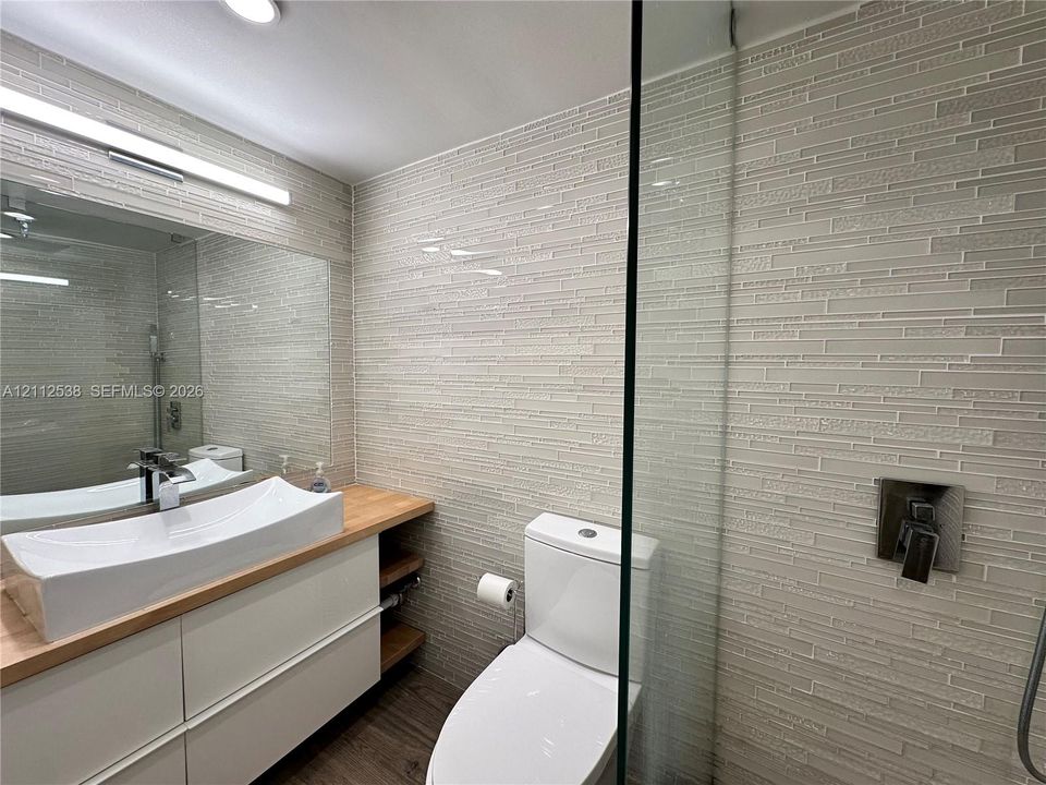Bathroom featuring a floating vanity with wood-tone countertop, rectangular vessel sink, wall-mounted faucet, full-wall horizontal glass tile, and a glass-enclosed shower with metallic hardware