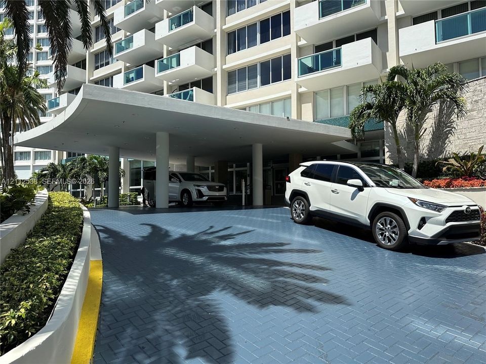 Covered porte-cochère entrance featuring a herringbone-patterned paved driveway, structural columns, and landscaped retaining walls