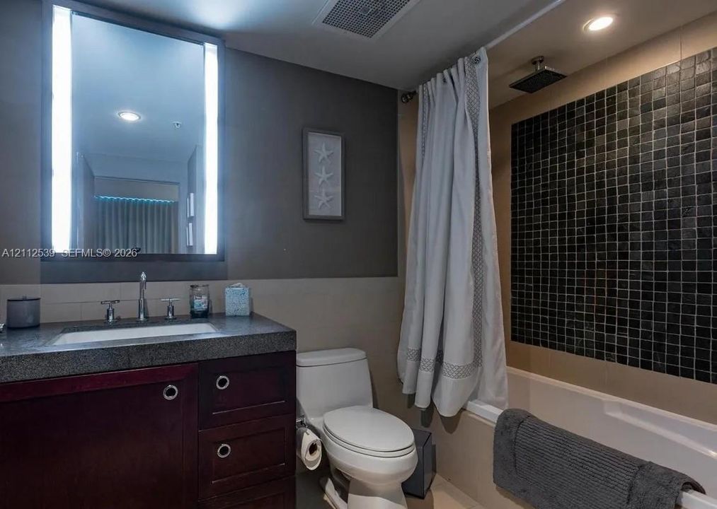 Bathroom featuring a dark wood-finish vanity with a stone countertop, an illuminated mirror, and a bathtub with black mosaic tile surround and rainfall shower head