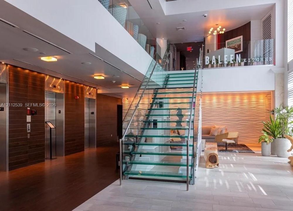Grand lobby entrance featuring a central glass-tread staircase with stainless steel railings, multi-level design, wood-paneled elevator surround, and textured wall accents