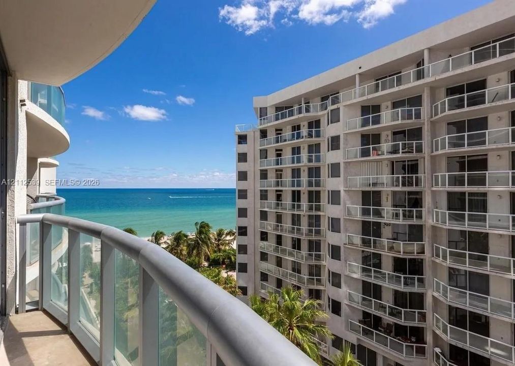 Private balcony featuring metal railings and glass panels, overlooking oceanfront scenery and coastal palm trees