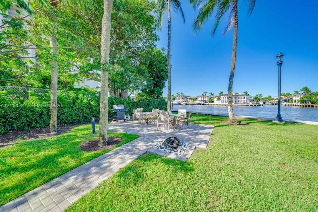 Waterfront grounds featuring a paver patio, manicured lawn, mature palm trees, and a decorative stone-lined fire pit area
