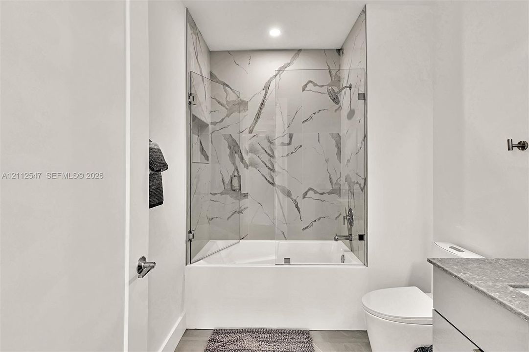 Full-height shower surround with veined tile, frameless glass enclosure, recessed shower niche, deep soaking tub, and stone-finish vanity countertop