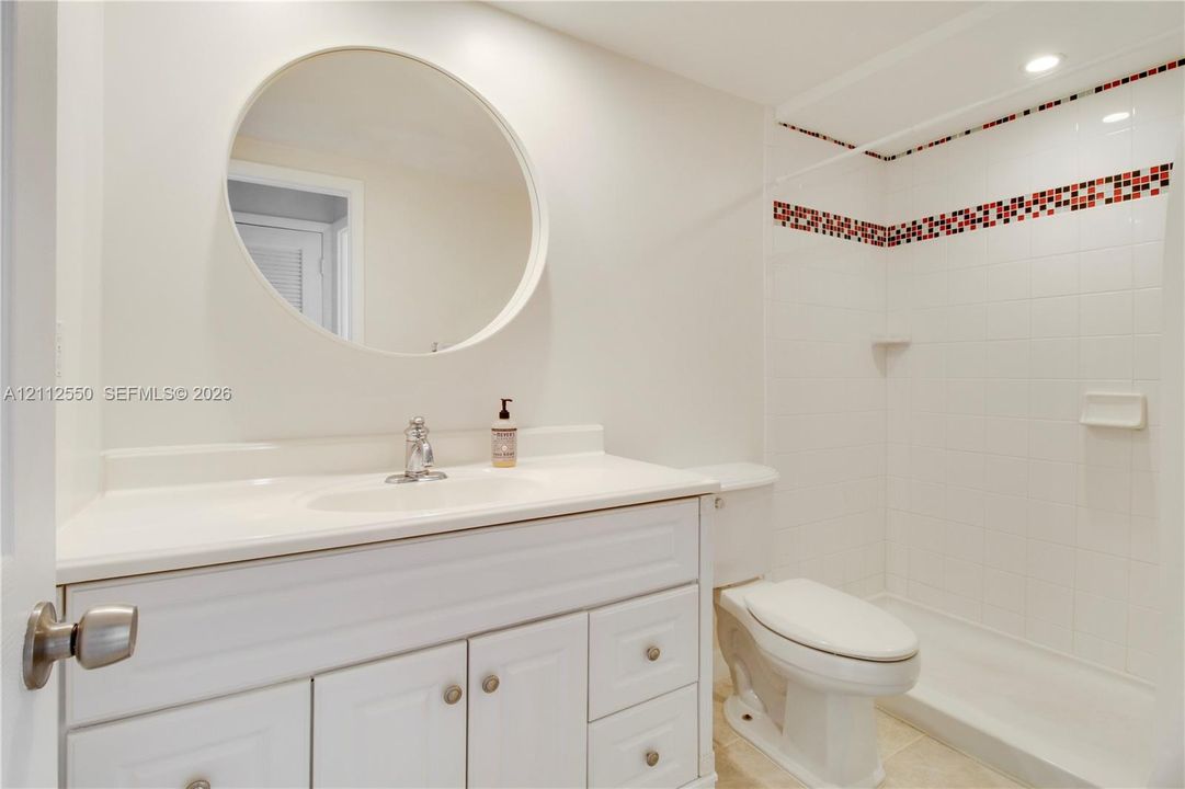 White vanity featuring a solid-surface countertop with an integrated basin, circular wall-mounted mirror, and a tile shower surround with decorative mosaic accents