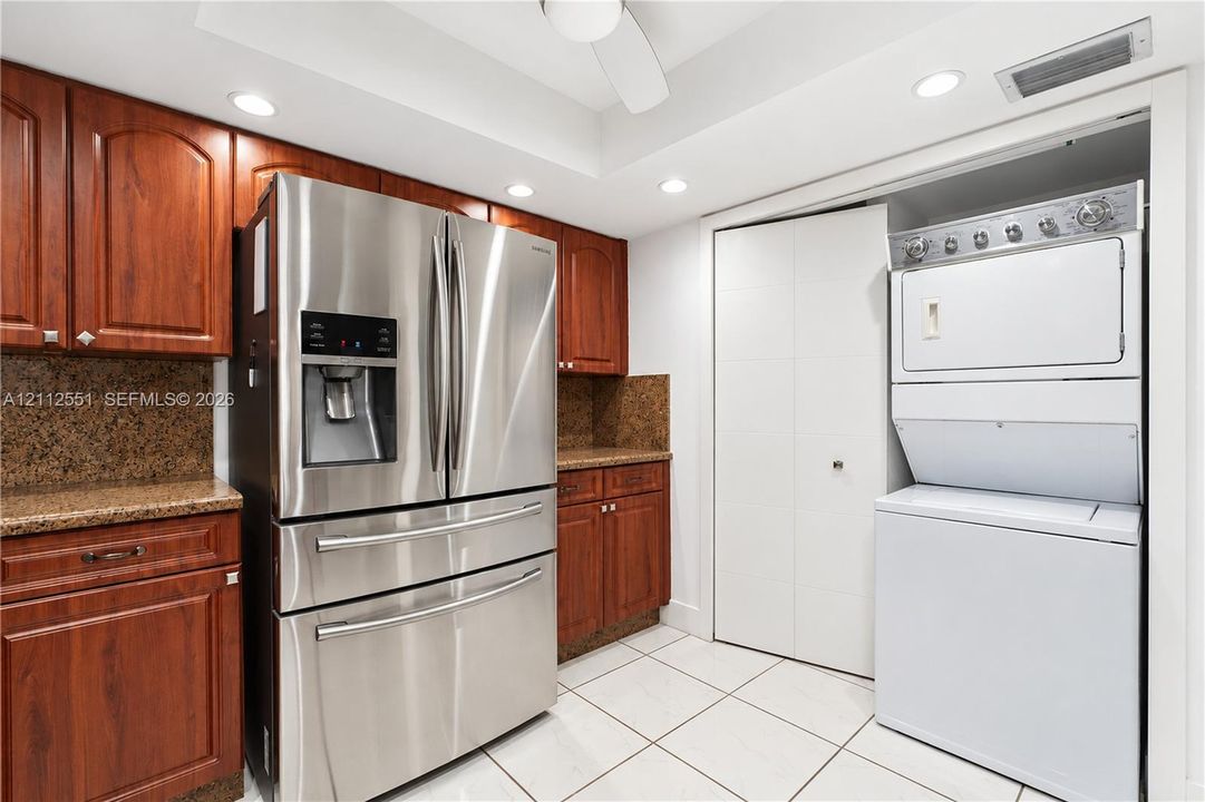 Wood-finish cabinetry with stone countertops, stainless steel French door refrigerator, stacked washer and dryer unit, and tile flooring