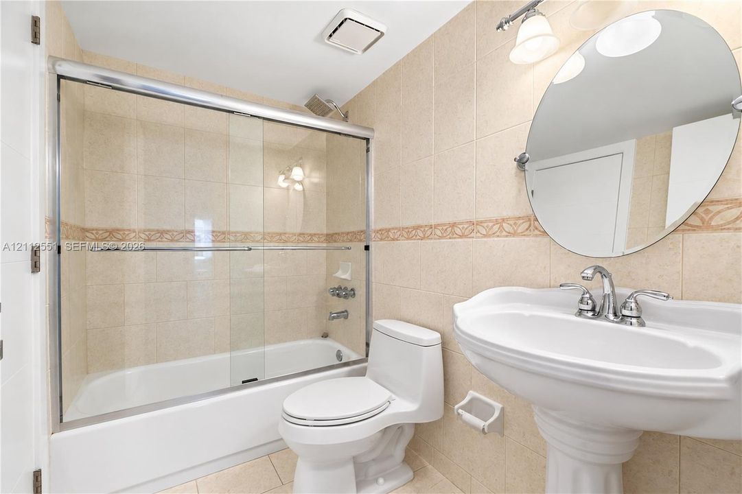 Bathroom featuring a pedestal sink with a chrome faucet, oval wall-mounted mirror, ceramic tile walls with decorative inlay, and a tub-shower combination with a glass sliding enclosure