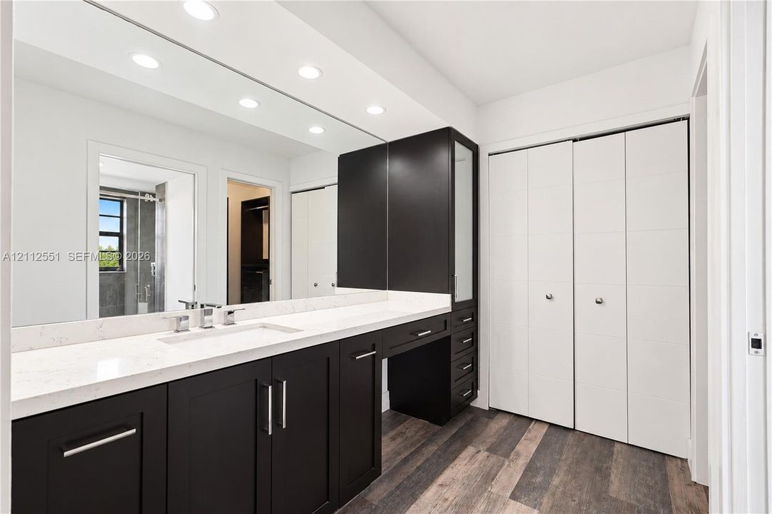 Dark wood-finish cabinetry, white stone-surfaced countertops, full-width wall mirror, recessed lighting, and integrated floor-to-ceiling storage with bifold doors