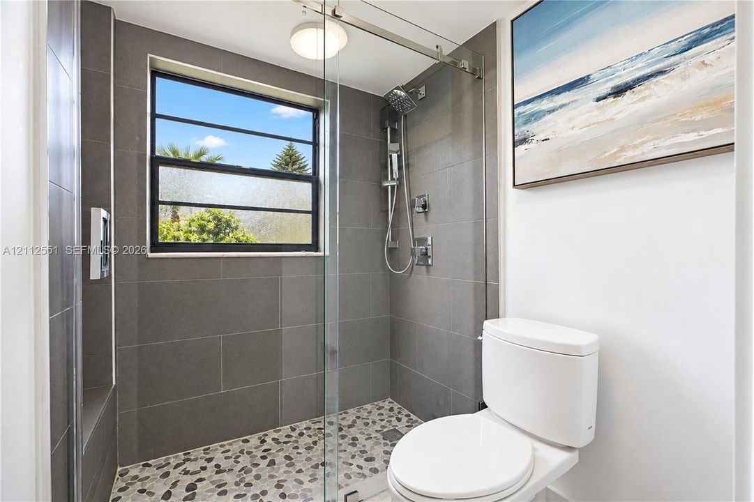 Walk-in shower featuring large-format grey wall tile, a pebble-stone floor, glass panel enclosure, and a chrome rainfall shower head