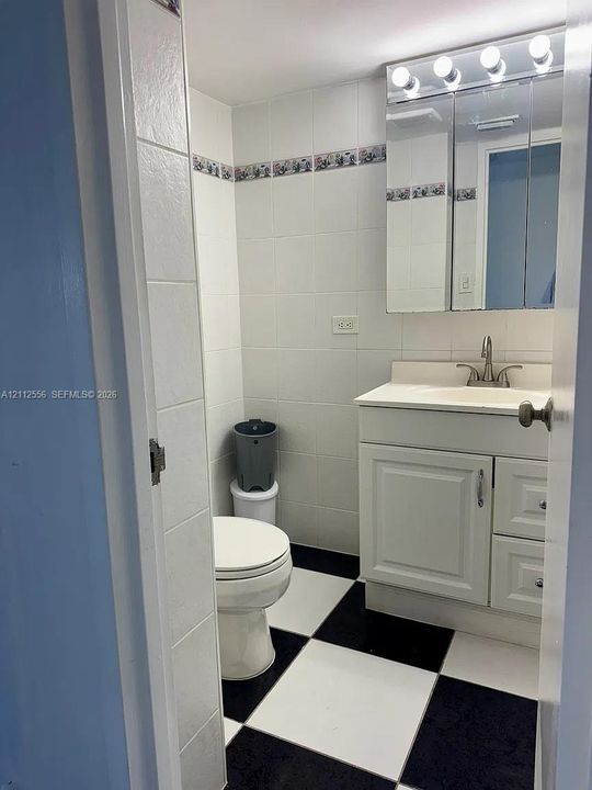 Bathroom featuring black-and-white checkered tile flooring, white wall tiling with decorative border, vanity cabinet with integrated sink, and mirrored medicine cabinet with overhead lighting