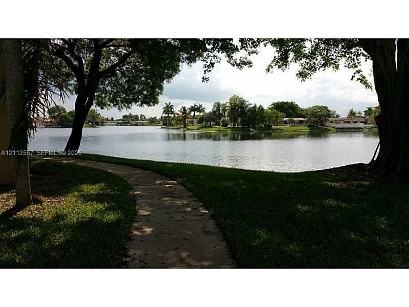 Waterfront grounds featuring a concrete walking path, mature trees, manicured turf, and open water scenery