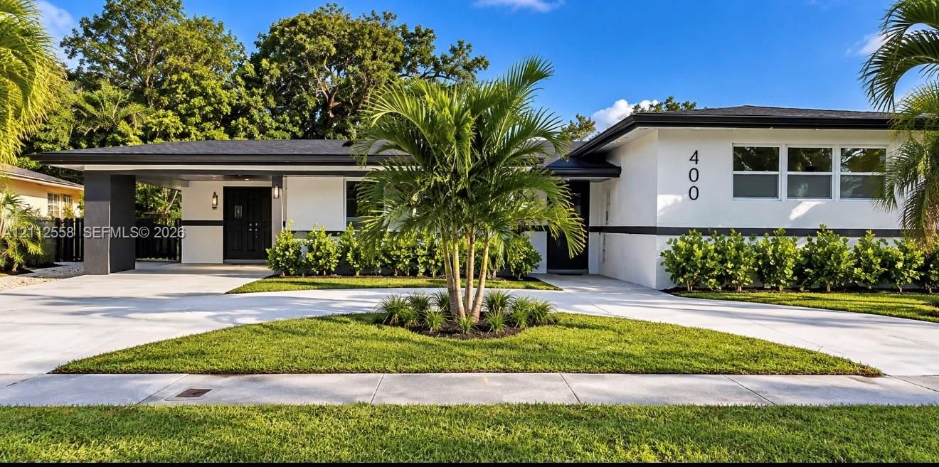 Mid-century exterior featuring a low-pitched roof, clean white stucco facade with dark trim, expansive concrete driveway, covered carport, and manicured landscaping