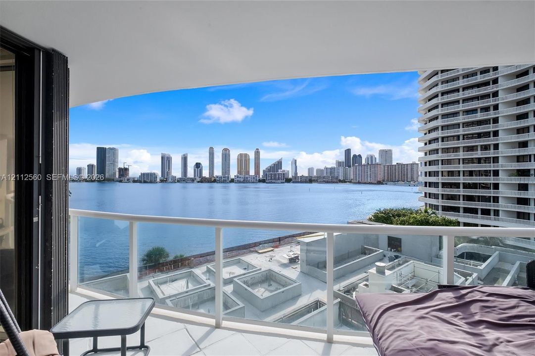 Private balcony featuring a glass-panel railing and tile flooring, oriented toward a water and city skyline backdrop