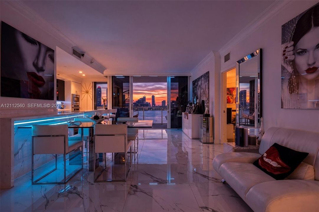 Open-concept living space featuring large-format polished stone-finish tile flooring, integrated linear LED accent lighting, and floor-to-ceiling sliding glass doors opening to a coastal cityscape and waterfront