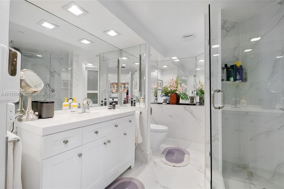 White vanity with an integrated countertop, full-width wall mirror, recessed ceiling lighting, and a glass-enclosed shower featuring large-format veined tile finishes