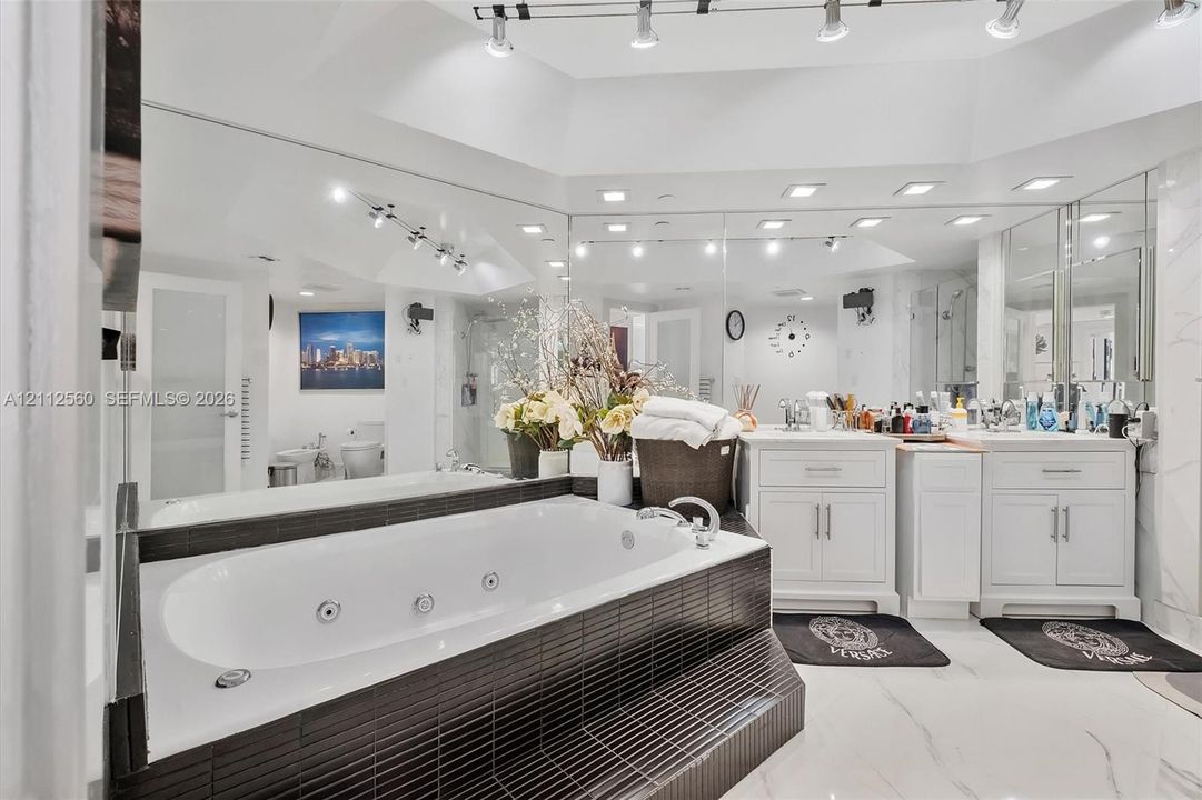 Jetted soaking tub with black tile surround, double vanity with white cabinetry, expansive wall-to-wall mirrors, recessed lighting, and large-format flooring