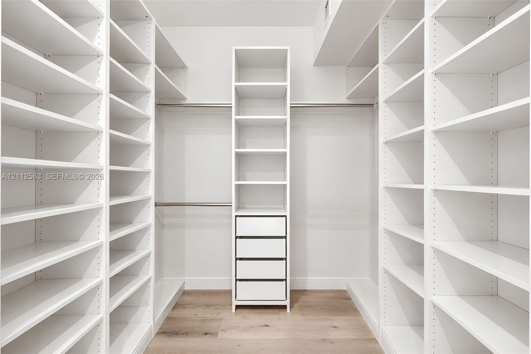 Walk-in closet featuring floor-to-ceiling white built-in shelving, integrated four-drawer stack, metal hanging rods, and wood-finish flooring
