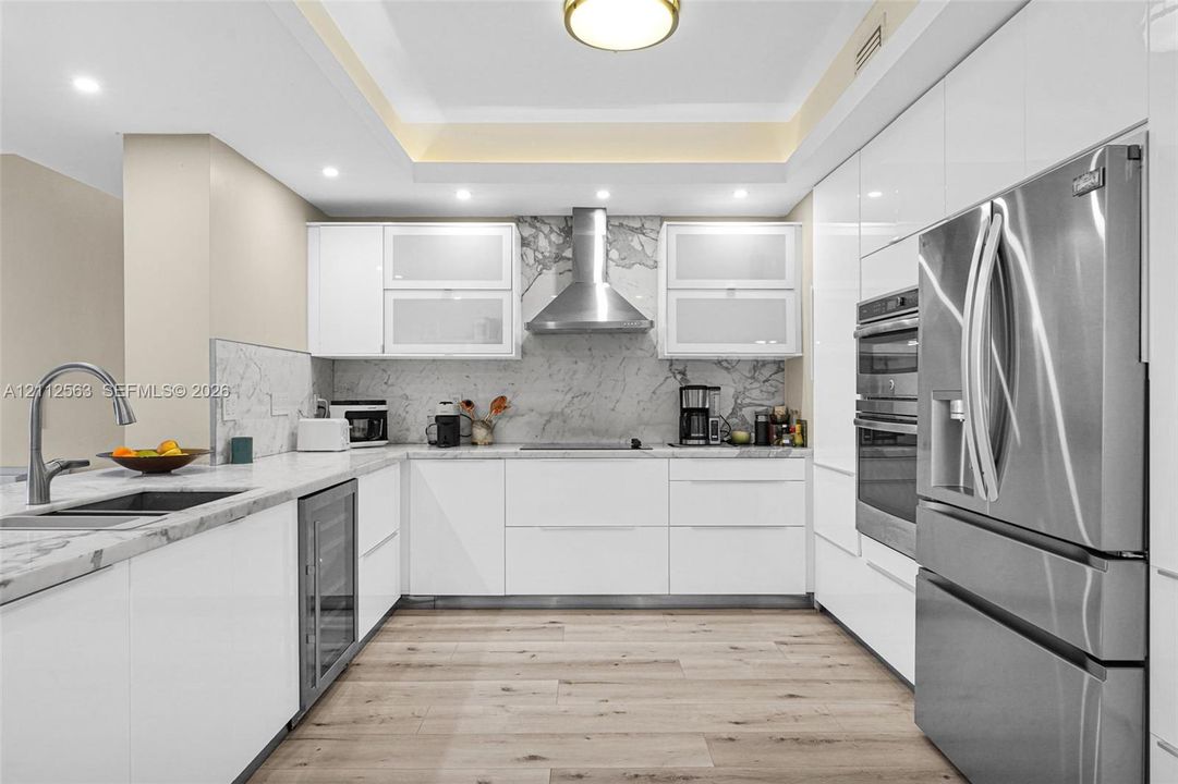 Contemporary kitchen featuring high-gloss white cabinetry, stone slab backsplash, stainless steel appliances, built-in dual ovens, and wood-finish flooring
