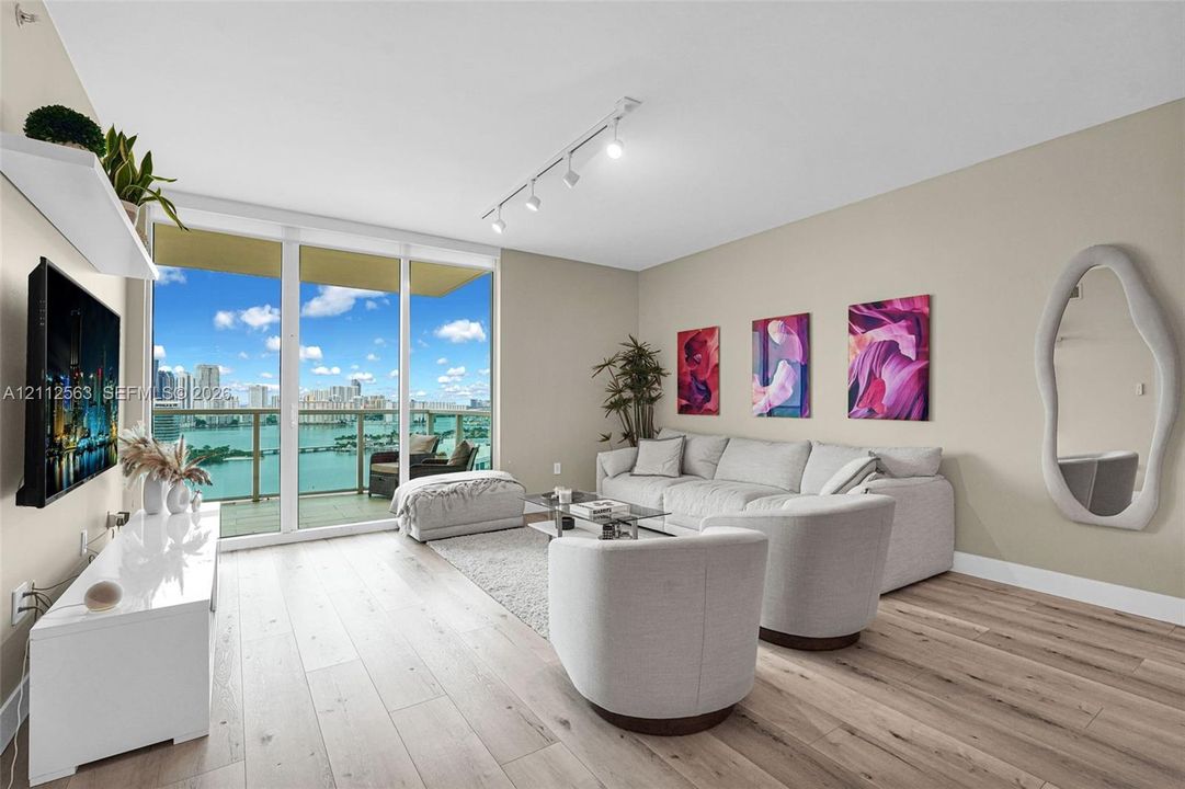 Floor-to-ceiling glass sliding doors, wood-finish flooring, recessed track lighting, and a private balcony featuring a water backdrop