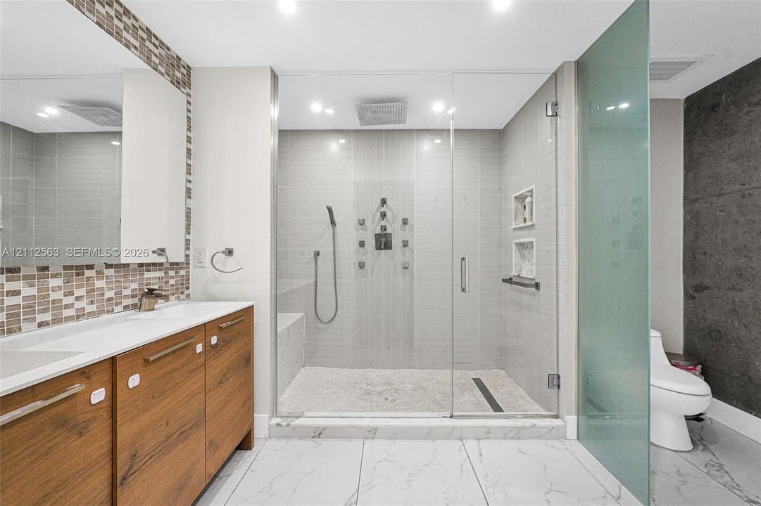 Bathroom featuring a floating wood-finish double vanity, walk-in glass-enclosed shower with multiple spray heads, built-in shelving, stone-finish flooring, and mosaic tile backsplash