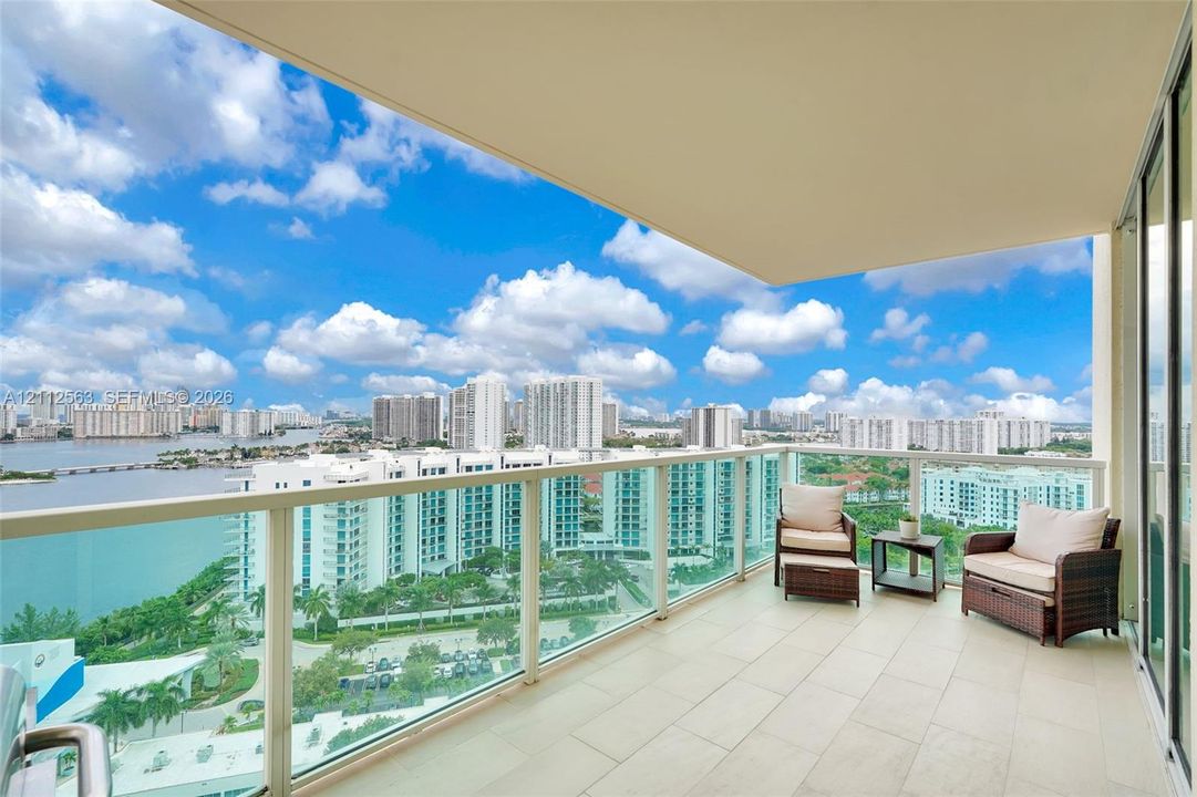 Covered balcony featuring neutral stone-finish flooring and a full-height glass railing, providing an unobstructed cityscape and waterfront panorama