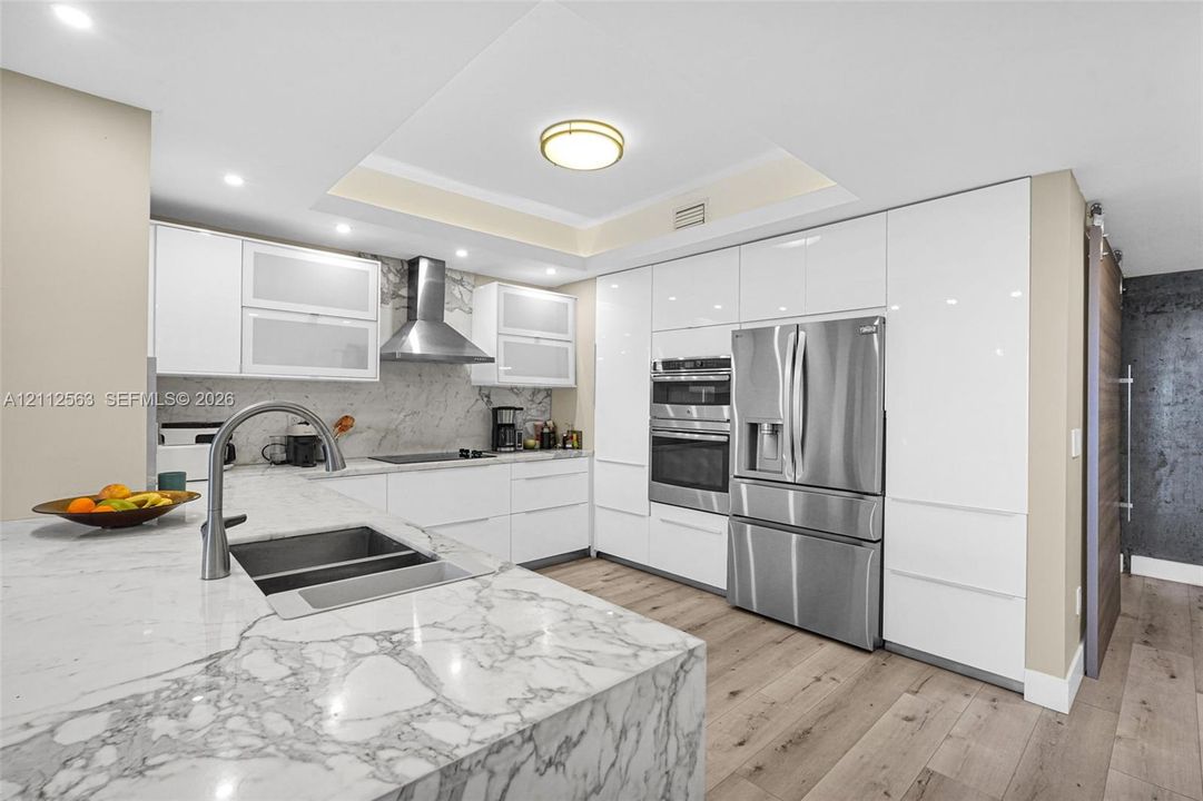 Modern kitchen featuring floor-to-ceiling high-gloss cabinetry, stone countertops, stainless steel appliance suite, wood-finish flooring, and a recessed ceiling design