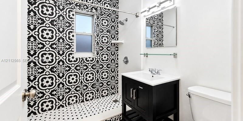 Bathroom featuring a black-and-white patterned tile shower surround, black vanity cabinet with an integrated white sink, rectangular wall mirror, and overhead vanity lighting