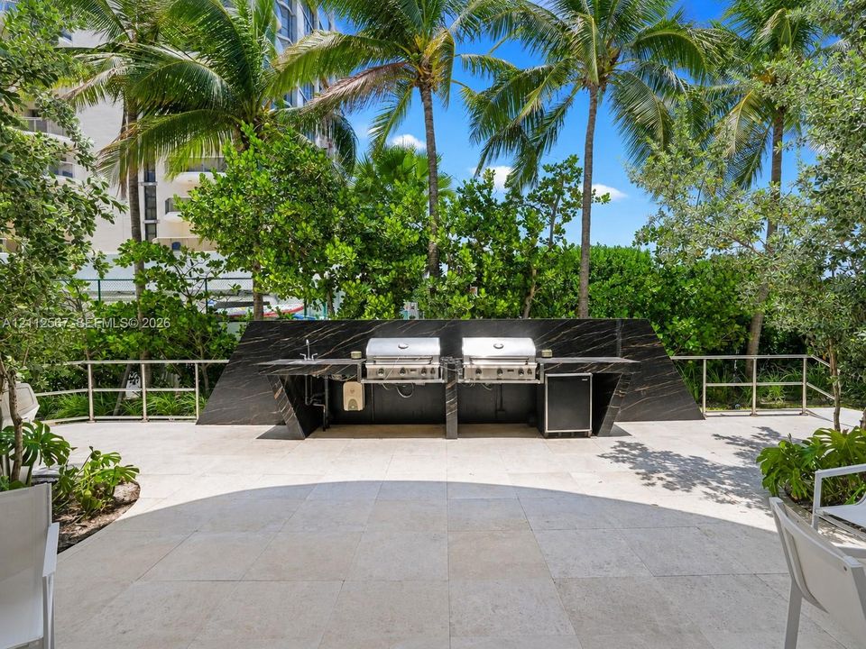 Outdoor kitchen featuring a dark stone-finish backsplash and countertop, stainless steel dual grills, integrated refrigeration, and a stone-tiled patio surrounded by mature palm trees