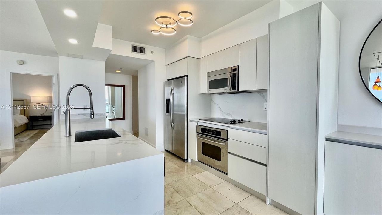 Kitchen featuring white flat-panel cabinetry, light-toned stone countertops, a waterfall-edge island, stainless steel appliances, and polished stone flooring