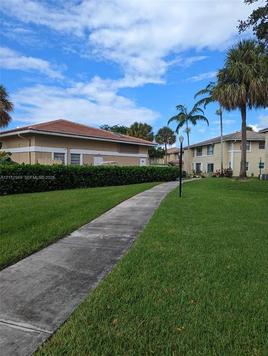 Concrete pedestrian path surrounded by landscaped grounds, manicured hedges, mature palm trees, and exterior pole lighting