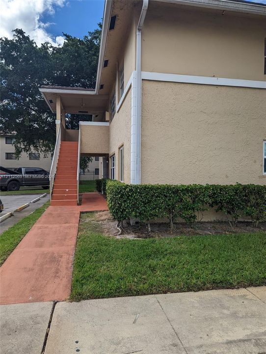 Stucco exterior featuring an elevated covered landing, integrated staircase, concrete walkway, and manicured hedge landscaping
