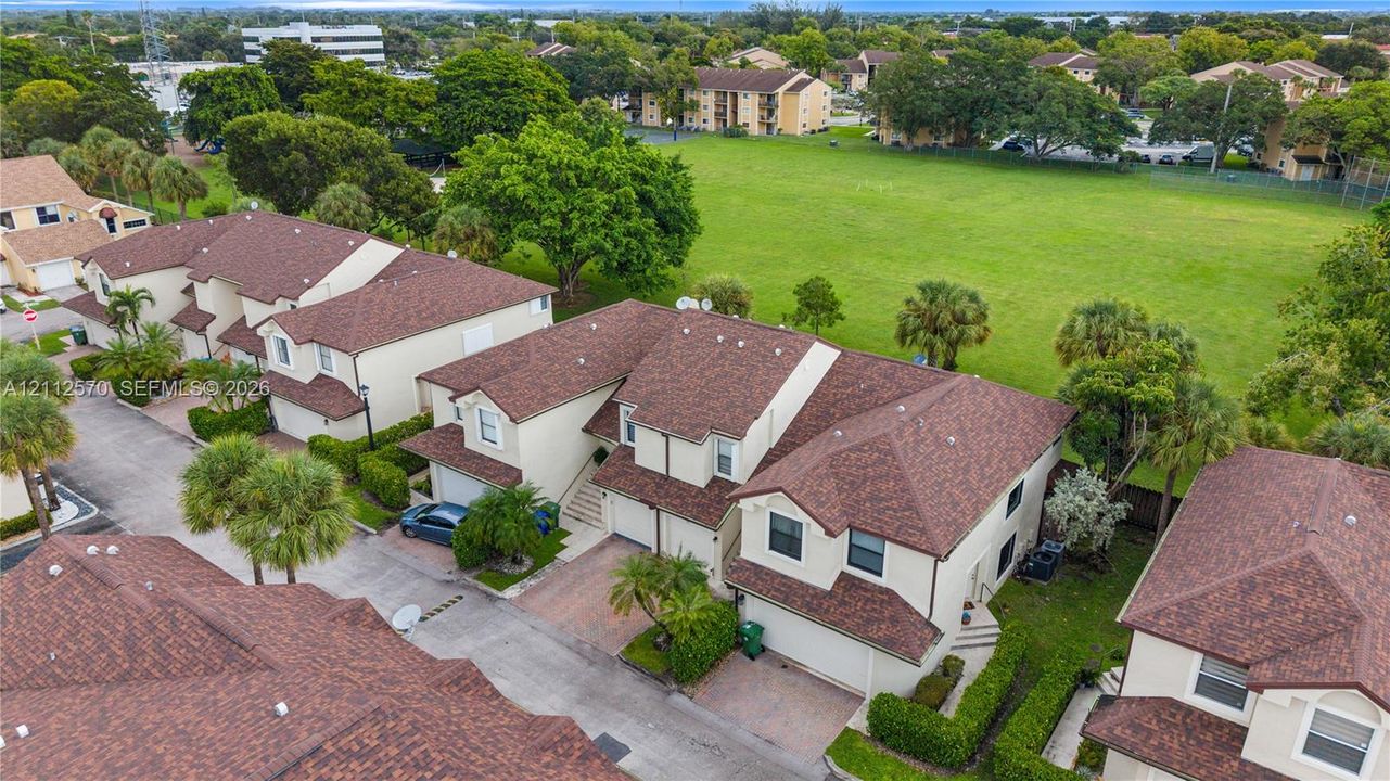 Aerial perspective of multi-story residences featuring shingled hip roofs, stucco facades, paved driveways, and integrated garages, situated amid landscaped grounds with palm trees