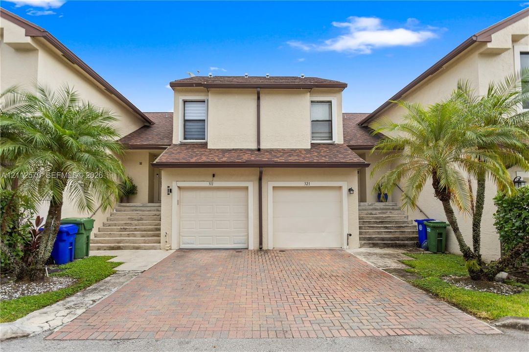 Stucco exterior featuring dual attached garages, a brick-paver driveway, shingle roofing, and concrete entry stairs