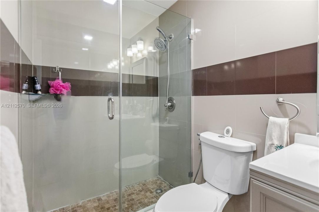 Walk-in shower featuring a glass enclosure, rainfall showerhead, and mosaic tile flooring, accented by dual-tone wall tiling and a neutral-toned vanity