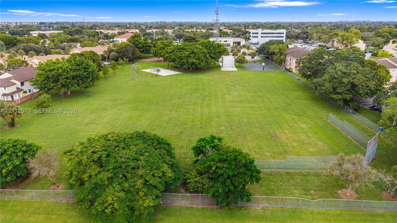 Expansive open green space featuring mature trees, chain-link fencing, a paved sports court, and a concrete recreational structure