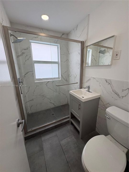 Walk-in shower with glass enclosure and large-format tile surround, grey vanity with integrated basin, matte finish floor tile, and recessed lighting