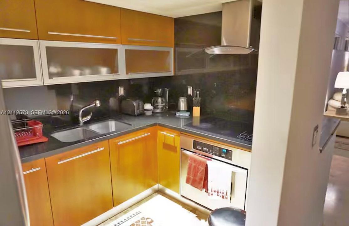 Kitchen featuring flat-panel wood-finish cabinetry, upper cabinets with glass-front inserts, solid-surface countertops, integrated stainless steel range hood, and stainless steel oven