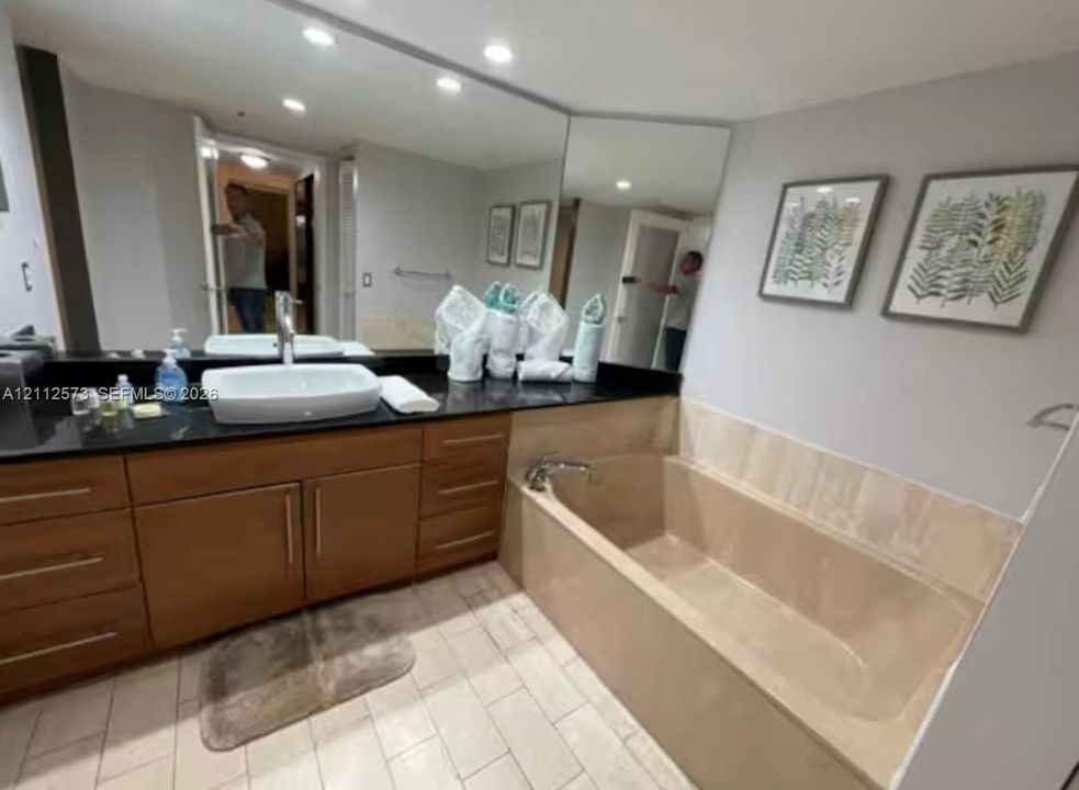 Wood-finish cabinetry with a dark countertop, white vessel sink, integrated wall-to-wall mirror, and a tile-surround soaking tub