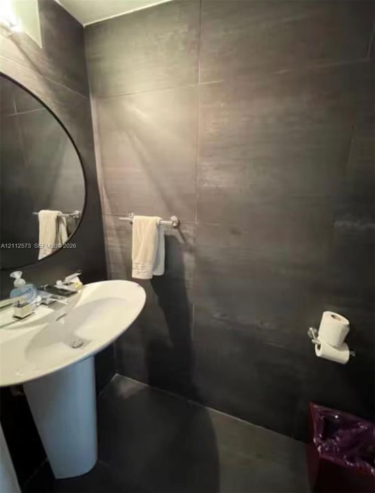 Large-format dark tile wall surfacing, circular wall-mounted mirror, white pedestal sink, and chrome fixtures