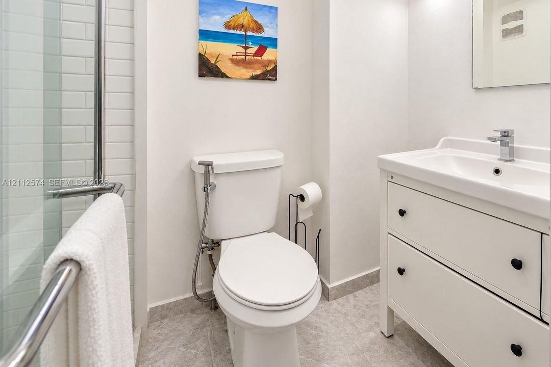 White vanity with integrated basin and matte hardware, glass-enclosed shower featuring white subway tile, and stone-finish flooring