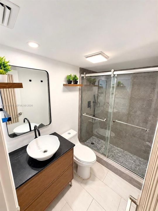 Modern bathroom featuring a wood-finish vanity with a black countertop, white vessel sink, matte black faucet, and a walk-in shower with stone tile surround, sliding glass door, and pebble tile flooring